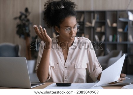 Serious Black woman reading and checking wrong financial papers, bills for paying with mistakes. Confused puzzled accountant reviewing tax documents for payment at workplace with laptop