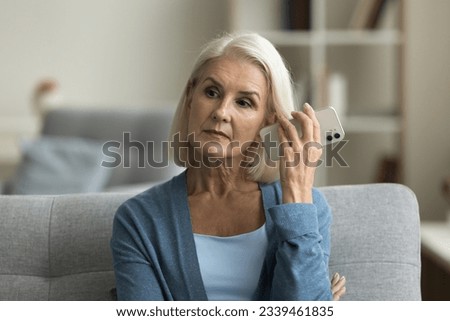 Serious blonde senior woman listening to voice message on smartphone, holding mobile phone dynamic at ear, looking away, thinking, sitting on home sofa, using Internet application, messenger on gadget