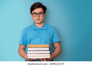 A serious boy, dark-haired, wearing glasses and a blue T-shirt, holding a stack of books - Powered by Shutterstock