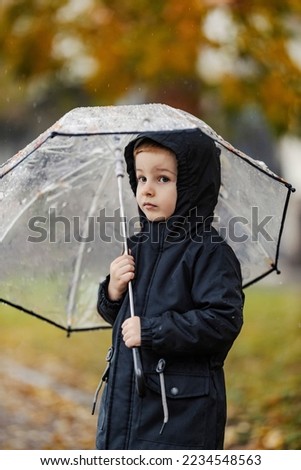 A serious boy is standing on the rainy weather on the street and hiding under umbrella.