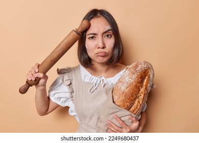 Serious brunette Asian woman busy baking bread at home holds freshly baked loaf and rolling pin smeared with flour looks tired wears apron stands against beige background. Bakehouse worker indoors - Shutterstock ID 2249680437