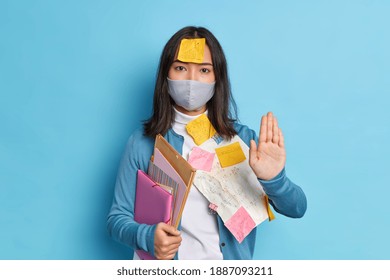 Serious brunette Asian woman office worker keeps palm towards camera in stop gesture asks not to bother her wears disposable mask as protection against coronavirus holds folders with documents - Powered by Shutterstock