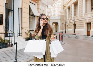 Serious Brunette Fashionista Lady Talking On Phone During Shopping In Autumn Weekend. Good-looking Shopaholic Woman Spending Time Outdoor And Making Purchases.