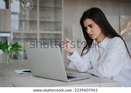 Serious brunette hispanic woman in white shirt sitting at desk with laptop makes video call disputes with colleague remote works at home. Beautiful female student talks via internet with parents.