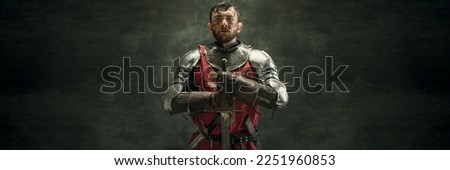 Serious brutal bearded man, medieval warrior and knight with dirty wounded face holding big sword over dark background. Concept of courage, history. Banner with copy space for ad