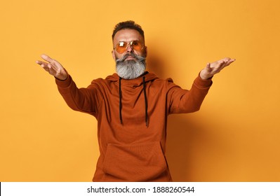 Serious brutal handsome grey-haired bearded man wearing casual sportswear, trendy sunglasses stretching hands to camera. Welcome, wide open hug. Indoor studio shot isolated on yellow background