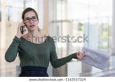Serious business expert concerned with latest news and making phone call. Young woman in glasses holding newspaper while talking on cell outdoors. Bad news concept