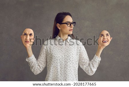Serious business lady holding two face masks of diametrically opposite joy sadness moods standing isolated on grey studio background. Person having extreme mood swing, bipolar disorder, schizophrenia