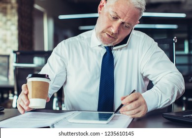 Serious Business Owner Negotiating On Phone. Mature Man In Tie Talking On Smartphone, Checking Data With Tablet And Holding Cup Of Coffee. Business Communication And Multitasking Concept