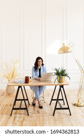 A Serious Business Working Freelance Girl A Woman With Black Hair In A Pale Blue Suit Working On A Laptop And Phone At A Wooden Table Against A White Wall In The Home Office