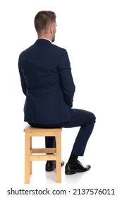 Serious Businessman In Elegant Navy Blue Suit Sitting On Wooden Chair In A Back View On White Background In Studio