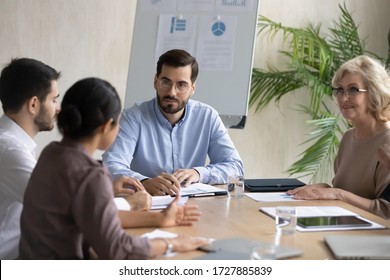 Serious Businessman Listening Indian Employee About Project Sit At Table In Boardroom At Company Meeting. Confident Leader Discuss Business Strategy With Diverse Woman And Colleagues.