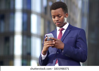 Serious businessman looking at his cell mobile phone, typing a message. Black African Afro American man in suit outdoors with smartphone. 