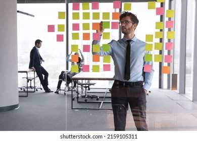 Serious businessman managing project tasks on sticky notes, writes start up business ideas using colorful post it stickers, plan corporate strategy on glass board. Creative priority to-do list concept
