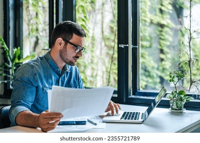 Serious businessman with papers using laptop at table. Young professional is analyzing strategy. He is working at table in home office. - Powered by Shutterstock