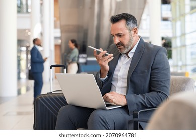 Serious businessman sitting on couch in airport lounge using laptop and sending voice message with smartphone. Mature business man working on computer while using voice command recorder on phone.