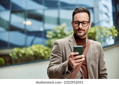 A serious businessman standing in front of the corporation and checking data on a smart phone. - Powered by Shutterstock