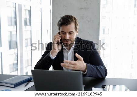 Serious businessman in suit working in modern skyscraper office, making call seated at desk looks at laptop talks to client on smartphone. Tech, workflow, lifestyle, distancing communication concept