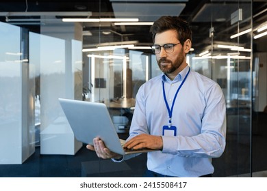 Serio hombre de negocios, administrador de sistemas parado con una laptop en las manos, un hombre pensante con una camisa lee los datos cuidadosamente, cerca de la ventana, un programador trabaja dentro de la oficina con una computadora.