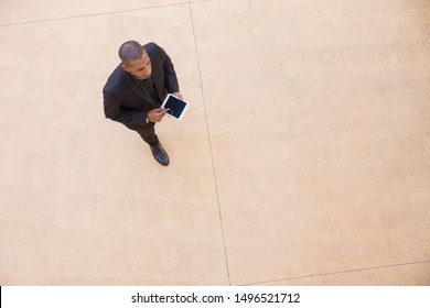 Serious Businessman With Tablet Going Through Office Hall. Top View Of Young African American Business Man Walking And Using Digital Device. Walking Businessman Concept