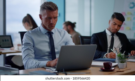 Serious Businessman Typing On Laptop In Open Space. Male Executive Using Laptop Computer In Office. Smiling Business Man Looking At Documents. Focused Man Making Notes In Notebook At Workplace