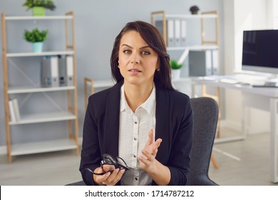 Serious Businesswoman In A Black Suit Looking At The Camera Talking To A Partner While Sitting In The Office.