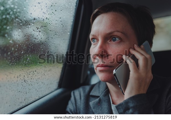 Serious businesswoman\
talking on mobile phone from the backseat of the car during the\
rain, selective focus