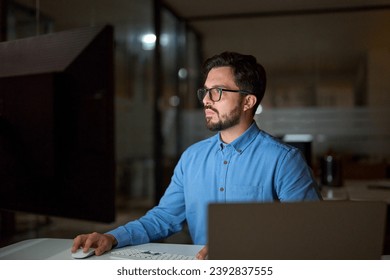 Serious busy concentrated young professional business man programmer software developer using desktop computer technology working online overtime late at night in dark office sitting at desk.