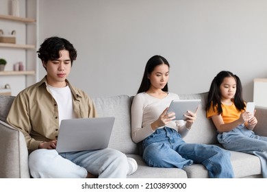 Serious Busy Japanese Millennial Mom, Dad And Teen Daughter With Personal Devices Sitting On Sofa In Living Room Interior. Work, Search, Browsing, New App, Online Game And Hobby At Home At Free Time