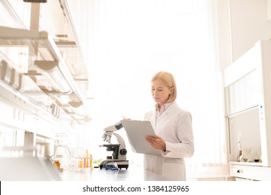 Serious busy mature female scientist in lab coat standing in laboratory room and checking information in clipboard before experiment - Powered by Shutterstock