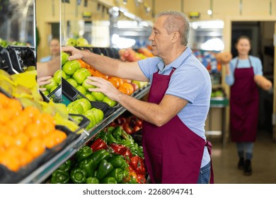 Serious busy mature male supermarket worker in apron arranging green apples in fruit section of store - Powered by Shutterstock