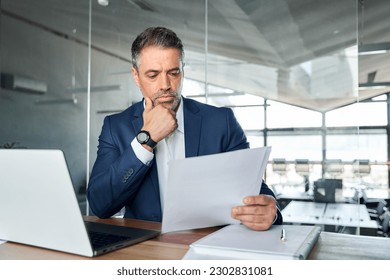 Serious busy mid aged professional business man lawyer or financial law expert wearing suit holding corporate documents reading paper contract sitting at desk in office managing risks feeling doubt. - Shutterstock ID 2302831081