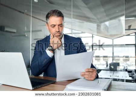 Serious busy mid aged professional business man lawyer or financial law expert wearing suit holding corporate documents reading paper contract sitting at desk in office managing risks feeling doubt.
