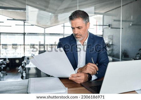 Serious busy middle aged professional business man executive ceo manager, lawyer or analyst wearing suit sitting at desk in office working checking bills corporate financial accounting documents.