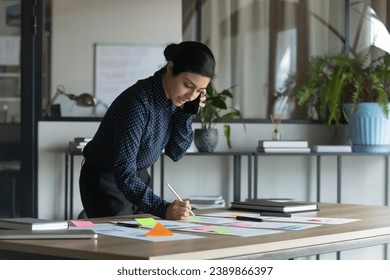 Serious busy multitasking young Indian businesswoman working with scrum arranged sticky papers, documents, graphic reports, writing notes, making call on cellphone, talking on mobile phone - Powered by Shutterstock