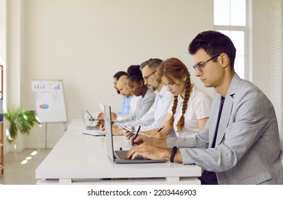 Serious Busy People Working In Modern Office Workplace. Group Of Diverse Employees Sitting In Row At White Desk, Working On Laptop Computers, Reading Business Reports, Taking Notes. Side Profile View