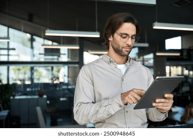 Serious busy young European business man entrepreneur using tablet standing in office at work. Male professional executive manager using tab computer managing financial banking data. Copy space. - Powered by Shutterstock