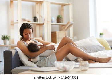 Serious calm careful young black mom sitting on sofa bed and breastfeeding baby while holding son in arms