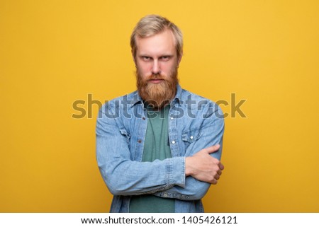 Serious and calm young hipster look scowl on camera. Angry and mad. Hands crossed. Wear jeans jacket and green shirt. Isolated alone on yellow background