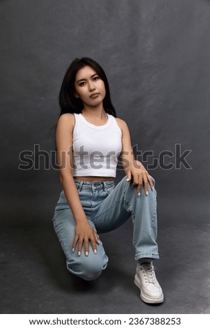Serious and calm young woman of Asian nationality sat down on one knee in fashionable jeans. Stylish young girl in modern clothes confidently holding on to the camera