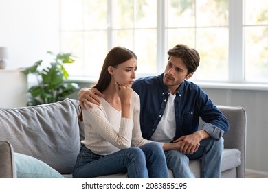 Serious Careful Young European Guy Calms Upset Unhappy Lady In Living Room Interior. Stress, Depression, Bad News, Family Support And Relationships Problems, Reconciliation After Quarrel, Infertility