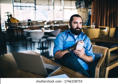 Serious Caucasian 40 Years Male In Casual Wear Working Remotely In Cafe Interior Using Smartphone And Laptop Computer For Research Online, Dreamy Mature Man Owner Of Small Business Planning Job