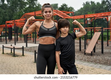 Serious Caucasian Athletic Mother And Son Showing Biceps And Looking At Camera On Sports Ground. Woman And Child Wearing Sportswear. Modern Healthy And Sports Lifestyle. Family Spending Time Together