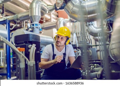 Serious caucasian handsome worker in overall, with hardhat and antiphons crouching and holding tablet while checking on motor. Factory interior.