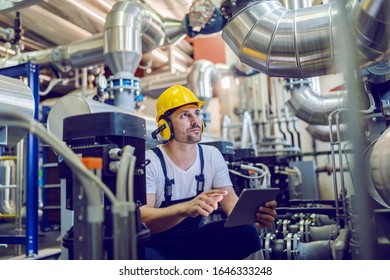 Serious caucasian handsome worker in overall, with hardhat and antiphons crouching and using tablet. Factory interior.