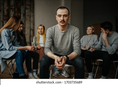 serious caucasian man, member of alcoholics anonymous club sit in center of isolated room. people support each other in the background, sit in the circle. help, support, problem, psychotherapy