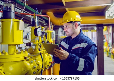 Serious caucasian unshaven worker in protective uniform   and hardhat using tablet for checking temperature in pipes  Factory interior 