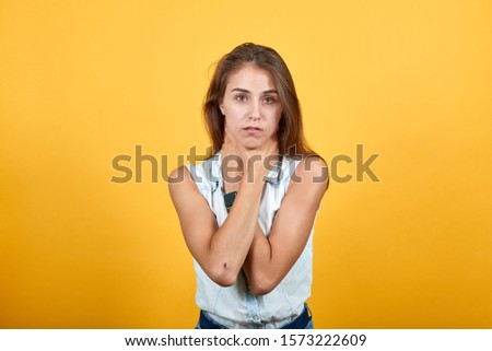 Serious caucasian young woman in blue denim shirt keeping hand on neck isolated on orange background in studio. People sincere emotions, lifestyle concept.