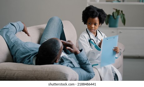 Serious Child Pretends To Be Doctor In Medical Costume Sitting Writing Symptoms On Tablet Father Lies On Couch Complains Of Feeling Unwell Painful Sensations African American Dad Playing With