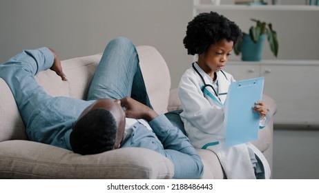 Serious Child Pretends To Be Doctor In Medical Costume Sitting Writing Symptoms On Tablet Father Lies On Couch Complains Of Feeling Unwell Painful Sensations African American Dad Playing With